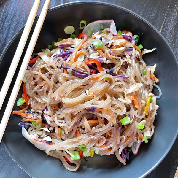 Glass noodles with shredded vegetables, including red cabbage, carrots, and bell peppers, in a black bowl with chopsticks.