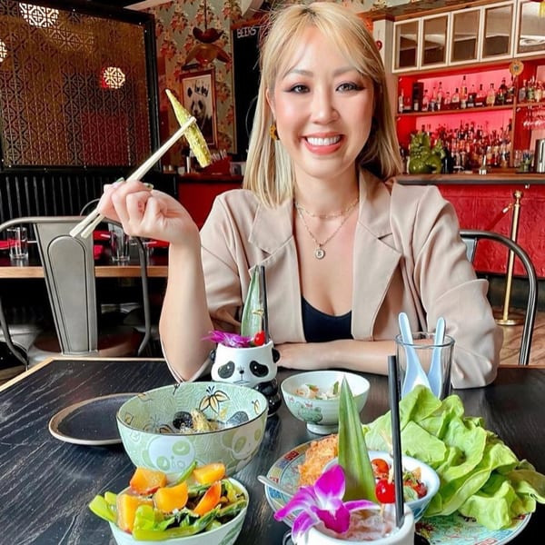 A smiling woman holds up a piece of food with chopsticks at Old Thousand, surrounded by Chinese dishes.