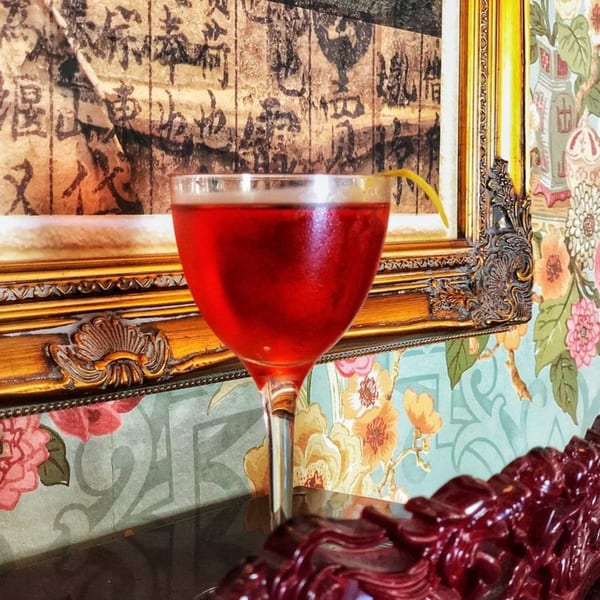A red cocktail with a lemon twist sits on a dark surface, framed by ornate decor and floral wallpaper.