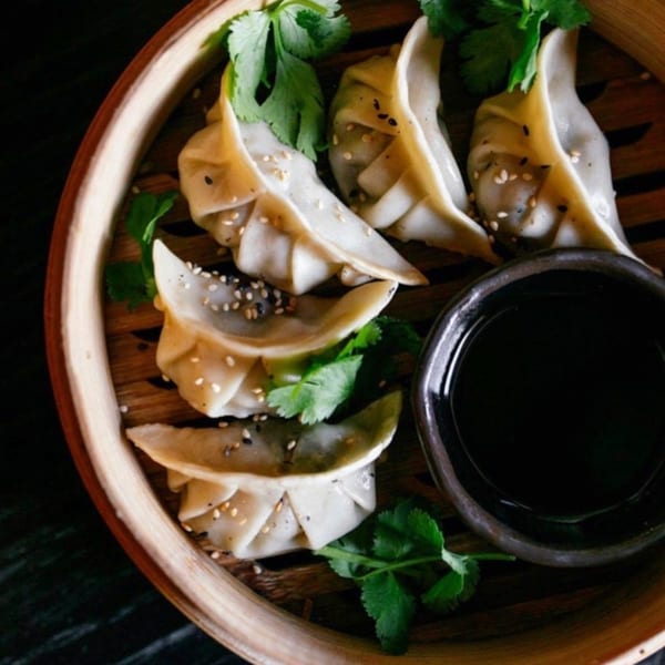 Steamed dumplings with sesame seeds and cilantro in a bamboo steamer with a side of soy sauce.