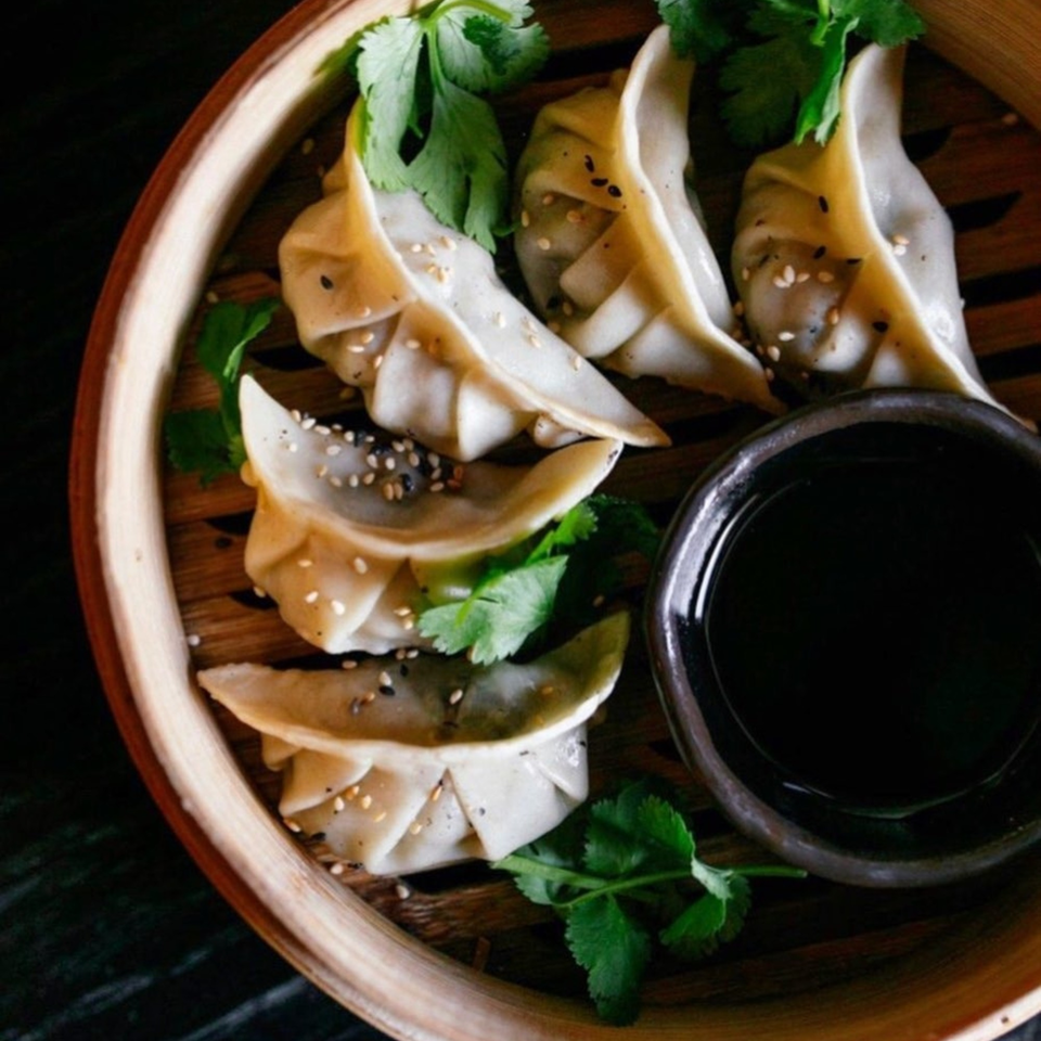 Steamed dumplings with sesame seeds and cilantro in a bamboo steamer with a side of soy sauce.