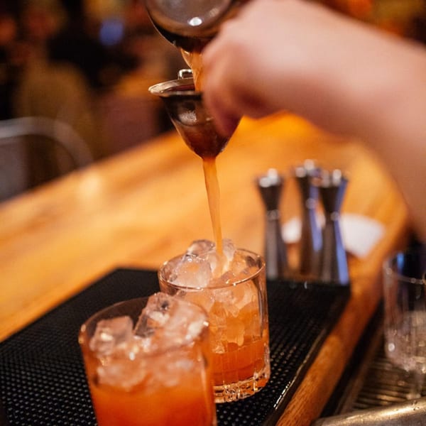 A bartender pours a vibrant orange cocktail from a shaker into a glass filled with ice.
