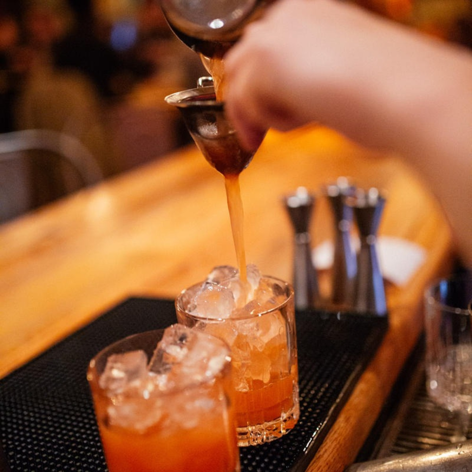 A bartender pours a vibrant orange cocktail from a shaker into a glass filled with ice.